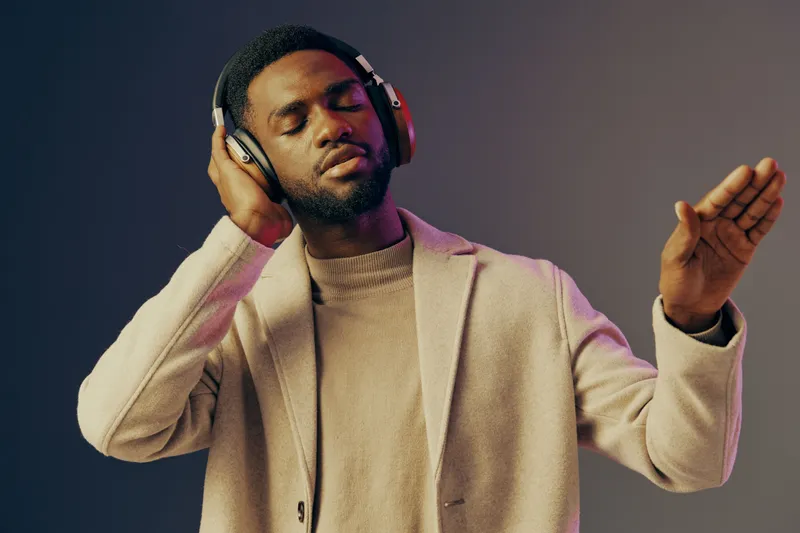 A stylish young Black man enjoying music with headphones, dressed in a light sweater and coat, expressing joy and relaxation in a modern, colorful background
