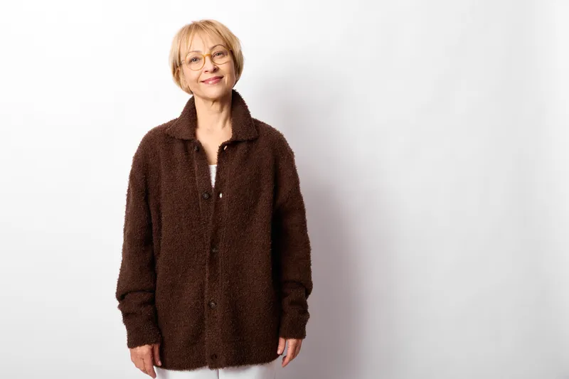 Elderly woman smiling in a studio portrait wearing a brown coat, standing on a white background, calm confident expression, casual winter fashion