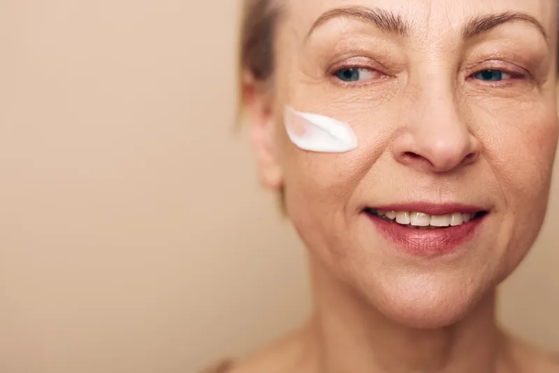 Smiling middle aged woman with natural skin applies white face cream on cheek, looking sideways, close up on beige background. Concept of skincare, aging, self care, beauty and wellness.