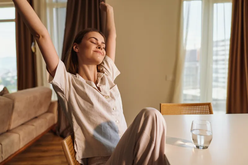 Woman in pajamas stretch at morning in a bright home, relaxed and smiling at a table with a glass of water, sunlight through the window in a cozy living room scene of calm wellbeing.