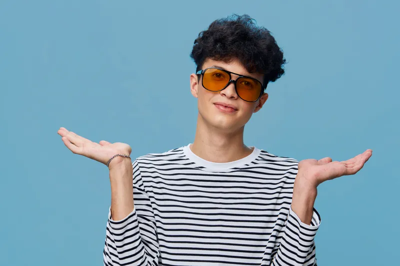 Young man with curly hair wearing striped shirt and amber sunglasses, posing with raised hands against a light blue background expressing casual uncertainty.