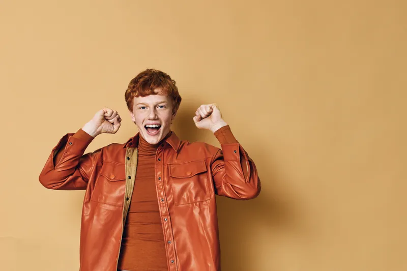 Boy with red hair and orange jacket celebrating with raised fists in front of plain beige background with happy excited facial expression.