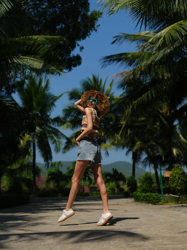 young woman with curly hair wearing sunglasses and denim shorts jumping on a sunny tropical path surrounded by palm trees and clear blue sky