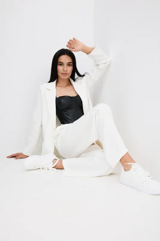 Young woman in white suit and black top poses stylishly against white minimalistic background, showcasing modern fashion with casual sneakers in bright setting.