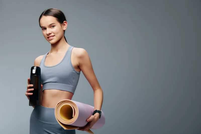 Young woman in athletic wear holding a hydration bottle and rolled yoga mat, ready for a workout. Fitness focused lifestyle, routine training, and calm studio backdrop convey motivation and active
