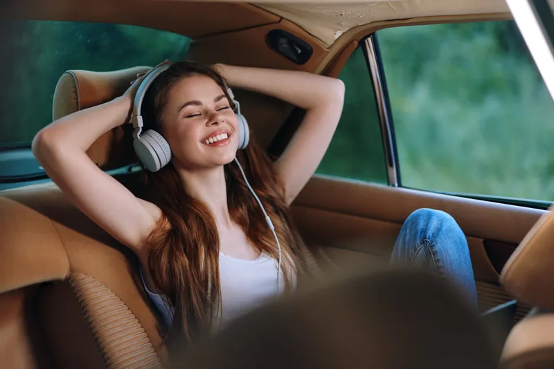 Happy young woman enjoying music with headphones in a car, relaxed and carefree during a sunny day, showcasing joy and serenity.