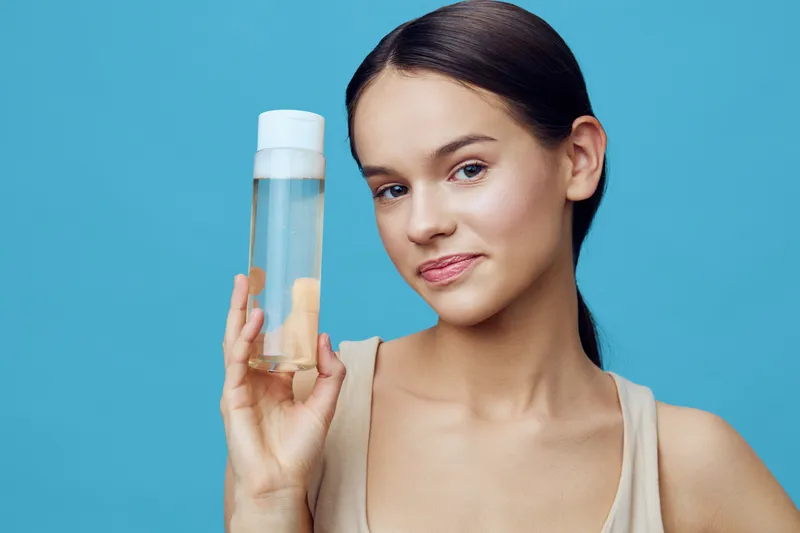 Young woman holding a skincare product, showcasing healthy skin with a confident smile against a blue background Ideal for beauty and skincare concepts