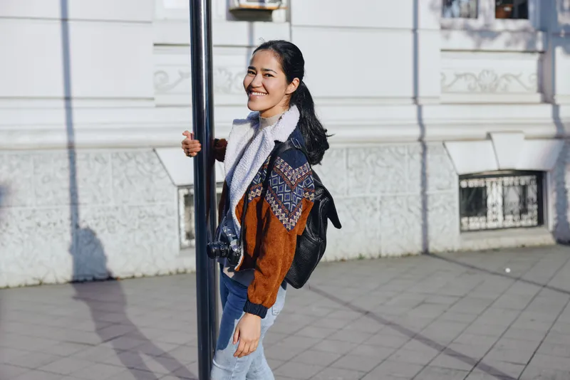 Woman walking outdoors along a city sidewalk with a scarf and backpack, smiling, wearing a brown jacket. Casual street style, urban vibe, daytime light, candid motion and energy.
