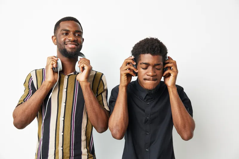 Two black men enjoying music together with headphones, showcasing happiness through their smiles and relaxed expressions in a minimalistic background.