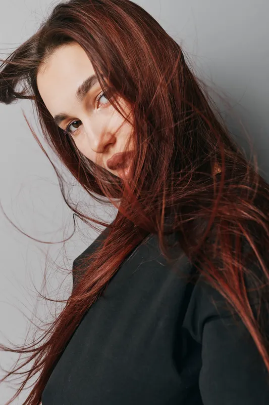 Woman with long red hair and natural makeup wearing black shirt posing with hair flowing in front of gray background for beauty portrait and fashion style.