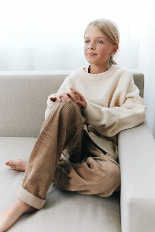 A young girl sits on a light sofa in a cozy home setting, wearing a soft beige sweater and corduroy pants, relaxed pose, calm expression, and gentle natural light.