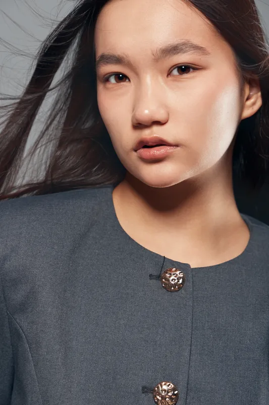 Young woman portrait with natural makeup and long hair wearing grey jacket with decorative buttons looking confident with neutral expression against plain background.