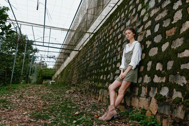 Young woman in casual outfit stands against a textured stone wall in a lush garden, conveying a sense of peace and harmony in nature