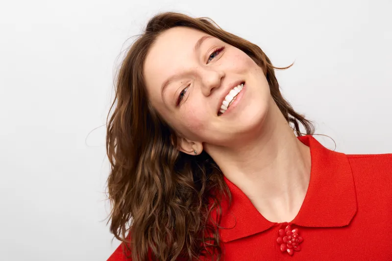 Smiling woman with wavy brown hair wears red textured sweater, tilting head slightly. Bright studio lighting highlights natural skin and joyful expression on white background.