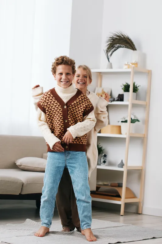 Two smiling children stand close indoors, the boy at front wearing a knit vest and jeans while a smiling adult hugs them from behind, creating a warm, lively scene in a bright living room with soft