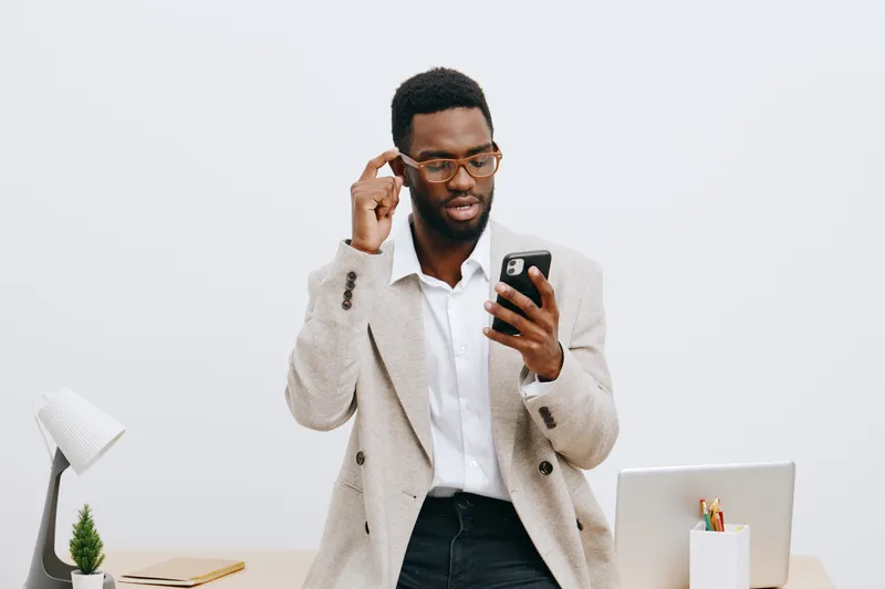 Modern young man with glasses checking smartphone in bright office, dressed in beige blazer and white shirt, freelance professional using mobile device, workplace environment, urban lifestyle.