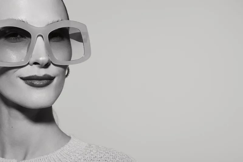 Portrait of a pale skinned white woman wearing large glasses with a confident smile on grey background.