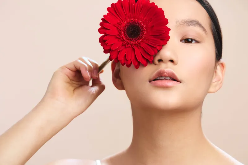 asian woman with a vivid red flower held near her face, soft light highlights smooth skin and calm expression, creating an artistic portrait with a clean neutral background