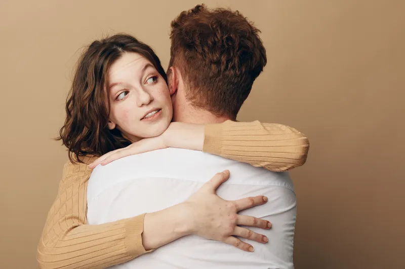 Young woman in beige sweater hugs man in white shirt, looking away with thoughtful expression. Intimate moment, relationship, affection, and emotional connection concept.