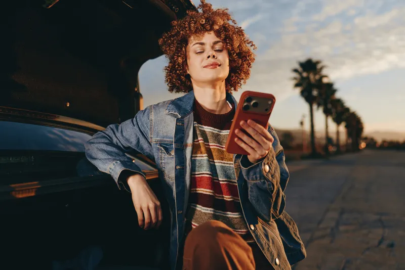 Curly haired woman lifestyle, wearing denim jacket, sitting by car at sunset on street, holding smartphone, smiling in warm evening light, casual travel moment