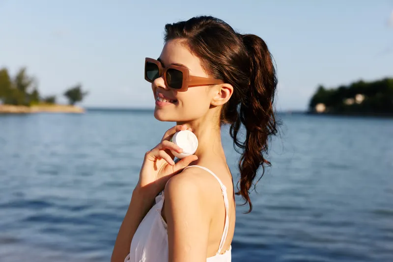 Young woman by a tranquil sea wearing sunglasses, holding a seashell, sunlit portrait, casual summer vibe, soft breeze, warm light, relaxed outdoor scene.