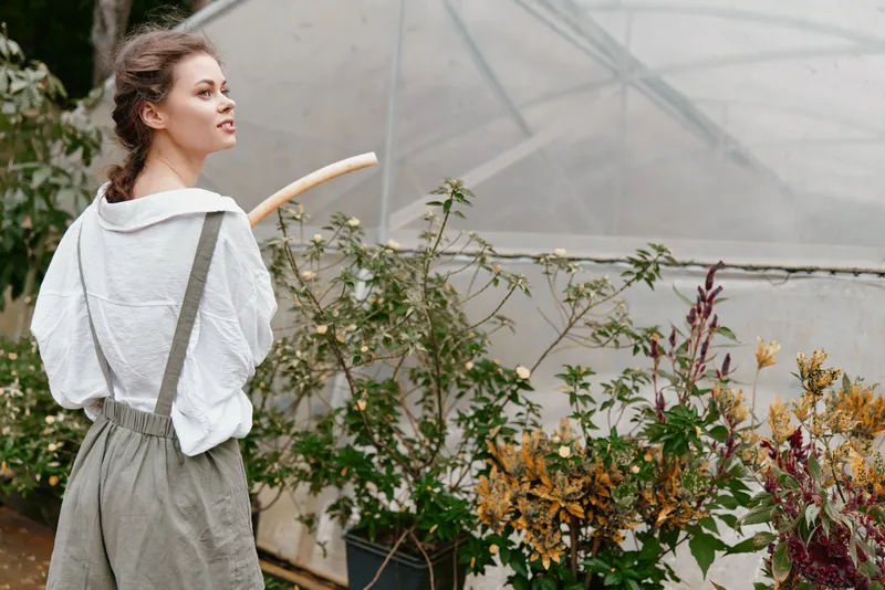 Young woman in overalls tending to plants in a greenhouse, showcasing a passion for gardening and connection to nature, dressed casually in earthy colors