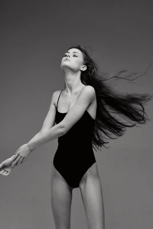 Black and white portrait of a young woman with flowing hair in a black swimsuit posing gracefully against a plain background.