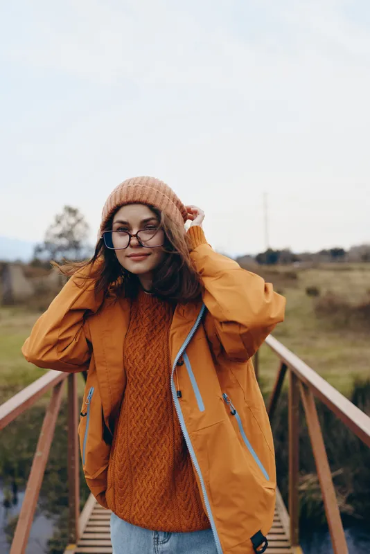 Woman in autumn lifestyle on an outdoor wooden path, walking, wearing orange coat, knit hat, sweater, and glasses, lifting hat with relaxed smile, soft daylight over open field, calm mood