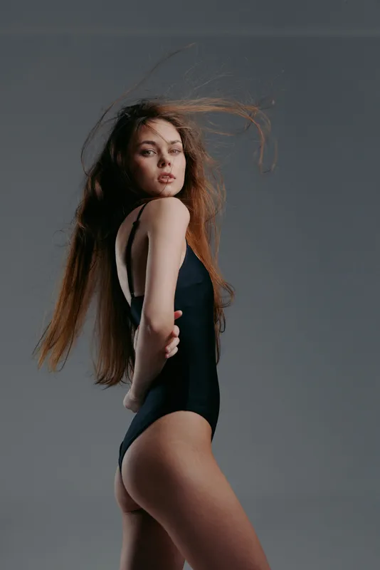 Young woman in black swimsuit posing confidently with flowing hair, studio background, minimalistic style, modern fashion portrait