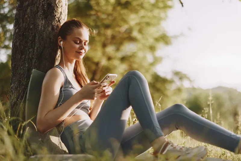 Young woman enjoying music with smartphone while sitting under tree in comfortable workout clothes, experiencing joy in nature during summer