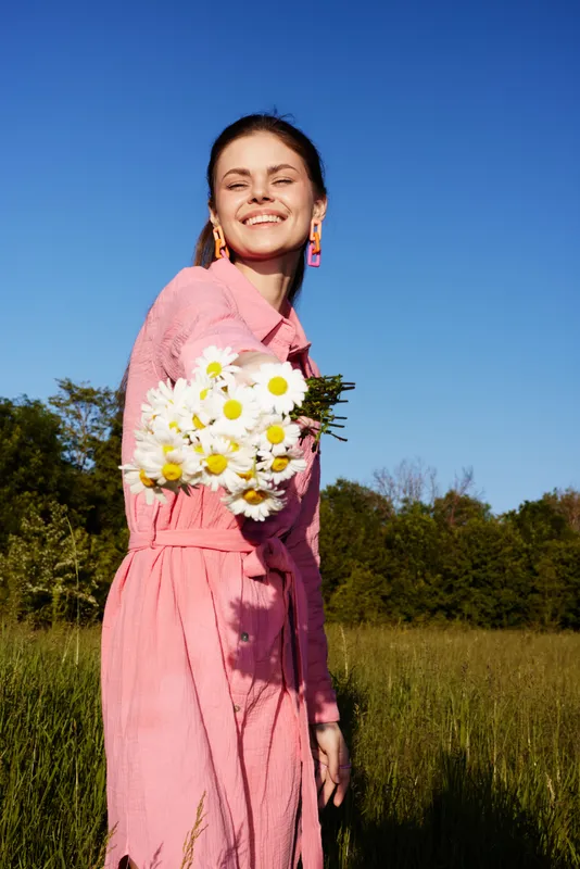 Young woman smiling joyfully in a pink dress, holding a bouquet of daisies in a sunny field The image captures a sense of happiness and nature