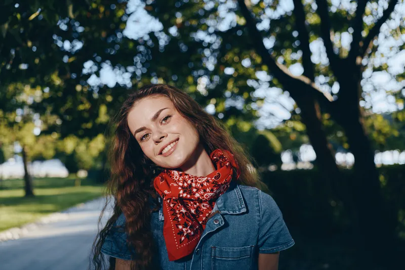 smiling woman with red scarf outdoors in summer park surrounded by trees and sunlight, casual style and natural beauty