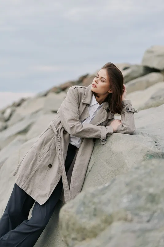 Young woman in beige coat leans on large rocks outdoors, looking away thoughtfully. Overcast sky and natural stone background create calm, contemplative mood in casual fashion portrait.