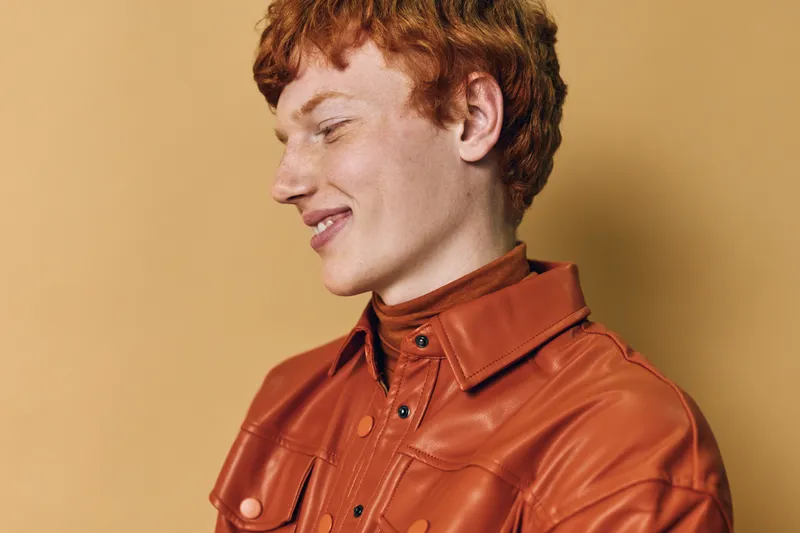 Young man with red hair and freckles smiling wearing orange leather jacket against neutral background. Casual portrait of confident male with natural expression and stylish clothing in studio.