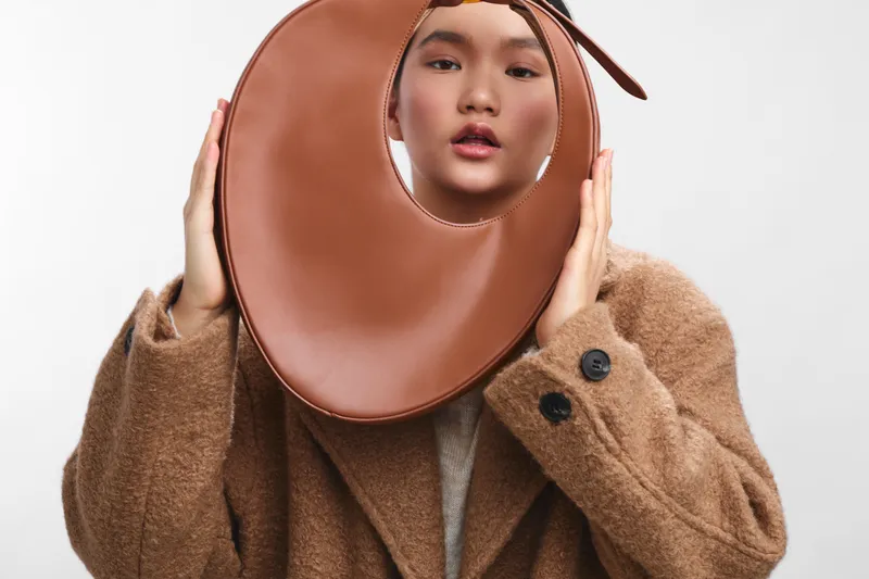 asian woman wearing a warm coat holds a large circular accessory near her face, creating a bold fashion portrait. The soft expression and minimal backdrop emphasize texture, color, and contemporary