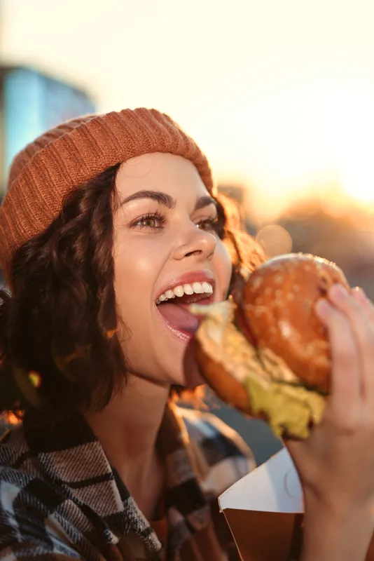 Woman eating burger with big smile in candid beanie outdoor lifestyle portrait during golden hour glow; authenticity and emotional storytelling capture mindful living and urban joy.