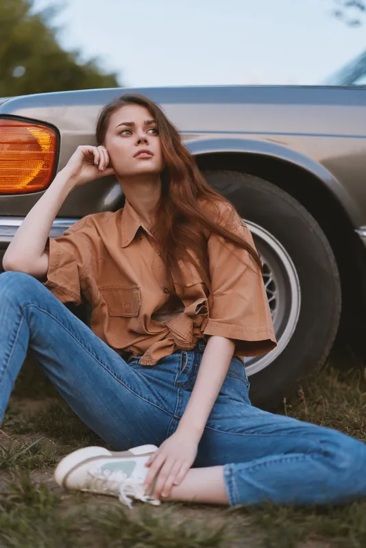 Young woman in casual outfit sits beside vintage car, showcasing laid-back style and thoughtful expression in an outdoor setting amidst greenery.