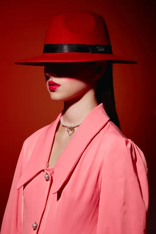 Woman in red hat and pink jacket poses against a red background. She wears bold red lipstick and a pearl necklace. Artistic creative fashion portrait with strong color contrast.