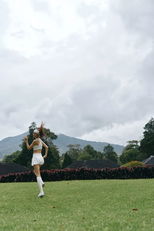 Young athletic woman jogging in a lush green field with mountains in the background, wearing a sporty outfit and expressing joy in a vibrant outdoor setting