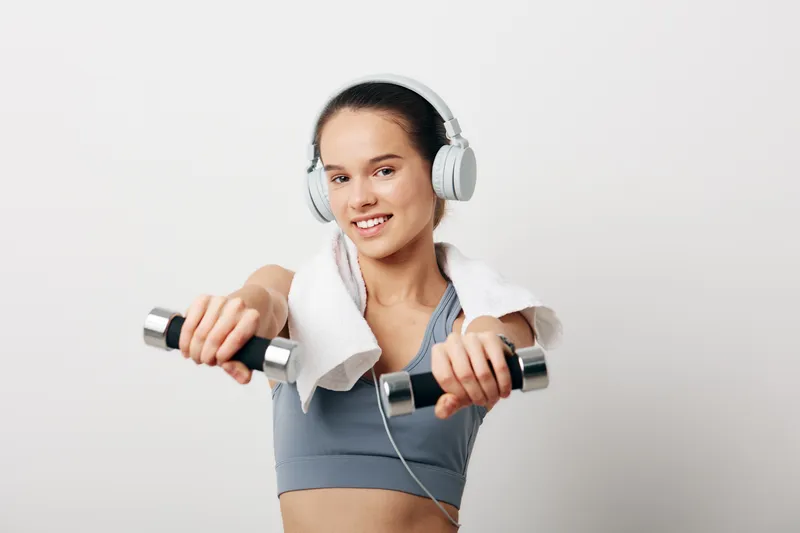 Young woman fitness portrait performing dumbbell workout, sport training in a sports bra with towel around neck and headphones, smiling confidently against a clean neutral studio background.