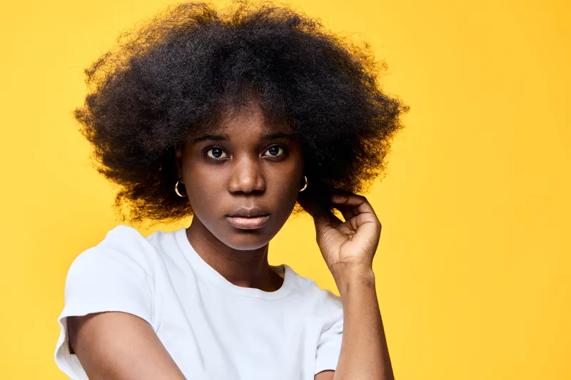 Young Black woman with natural hair in a white shirt, posing thoughtfully against a bright yellow background, expressing confidence and beauty
