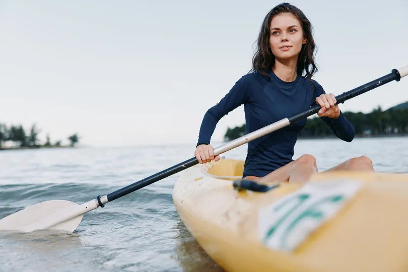 A focused young woman paddles a kayak on calm water, gripping the paddle with effort and balance as she glides toward the shore under soft daylight.