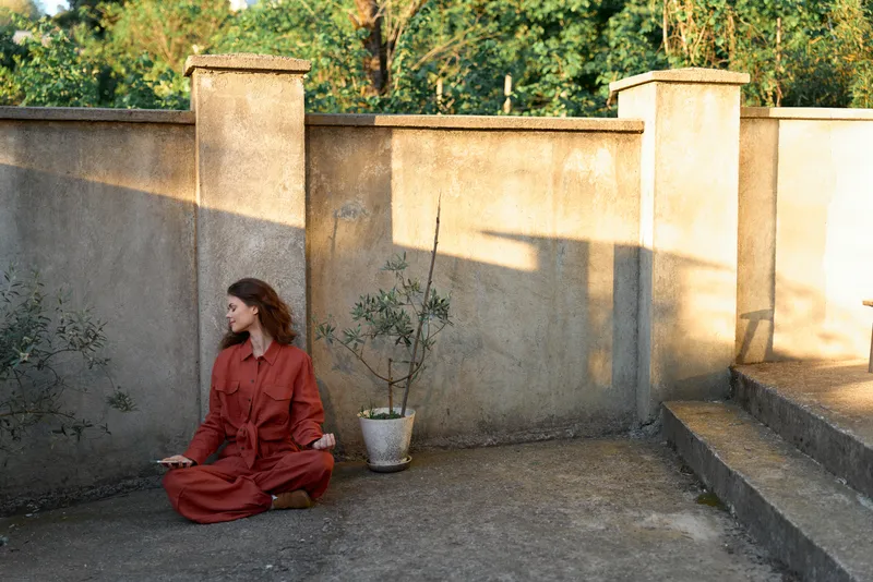 Woman wearing a red dress sits cross legged on a concrete terrace beside a potted plant, bathed in natural sunlight, exuding a calm outdoor lifestyle moment with a minimalist setting and warm mood