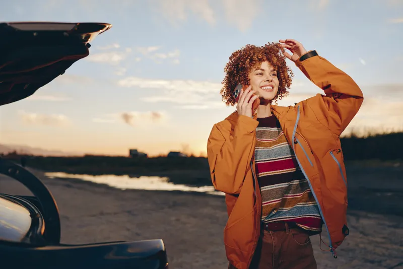 Woman lifestyle, curly hair, orange jacket, outdoors near a car at sunset, smiling and touching her hair, relaxed casual road trip mood