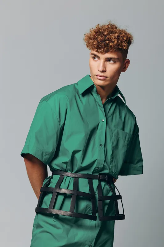 Young man with curly hair wearing green shirt and pants posing with black cage belt accessory on gray background. Fashion model looking sideways with serious expression in studio lighting.