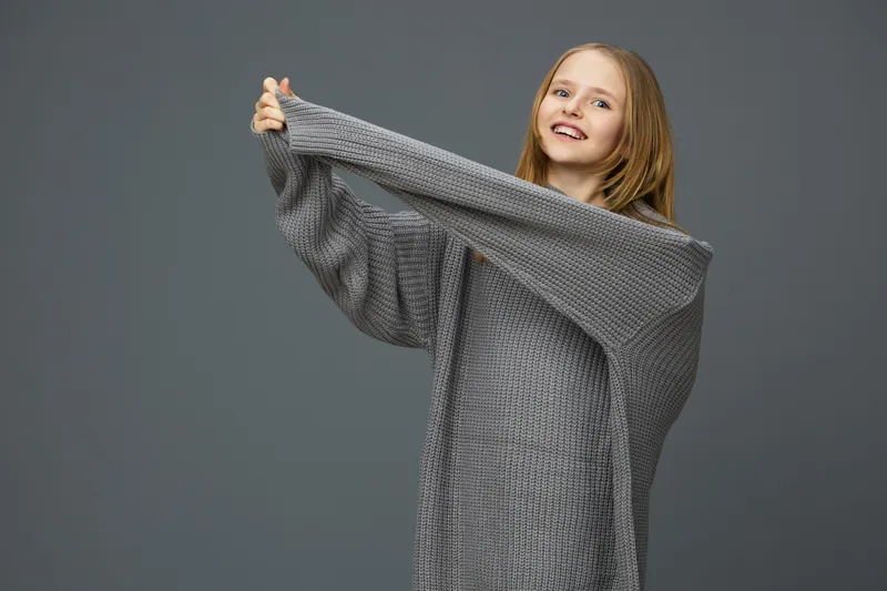 Young girl smiling in oversized gray sweater, showcasing cozy fashion against a muted background, representing comfort and modern style