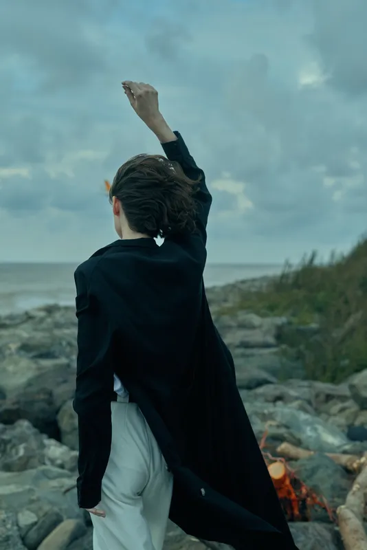 Woman stands on a rocky coast, wind in a dark jacket, raising a hand toward the sky as sea and clouds meet the horizon in a dramatic outdoor scene.