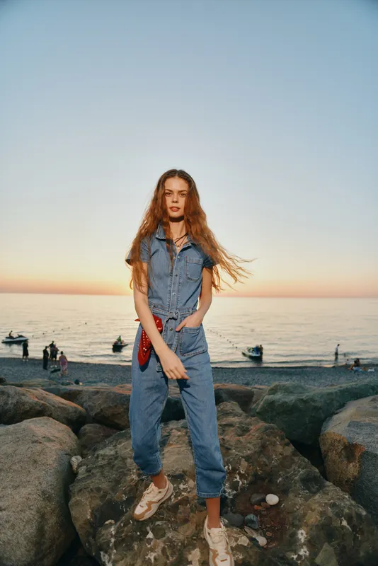 Lifestyle woman in modern boho-western denim street style stands on rocks by sunset sea, featuring film color tones and casual summer outfit with long hair flowing.
