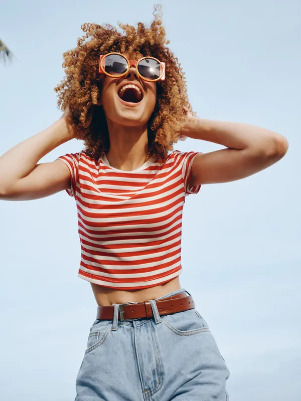 Joyful young woman with curly hair wears a striped crop top and jeans, laughing with sunglasses against a clear blue sky, capturing carefree summer energy.