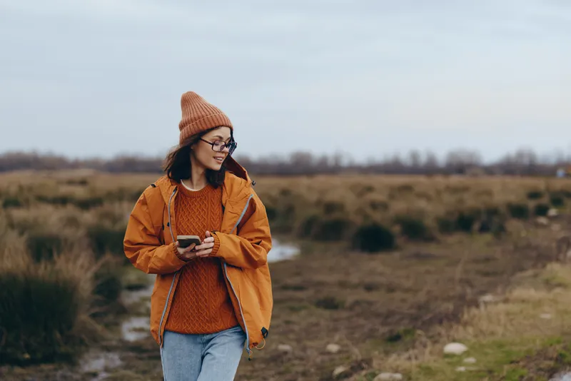 Woman lifestyle outdoors, countryside field, orange jacket and knit beanie, scarf near chin, phone in hands, smiling during cool winter walk, overcast sky, calm nature